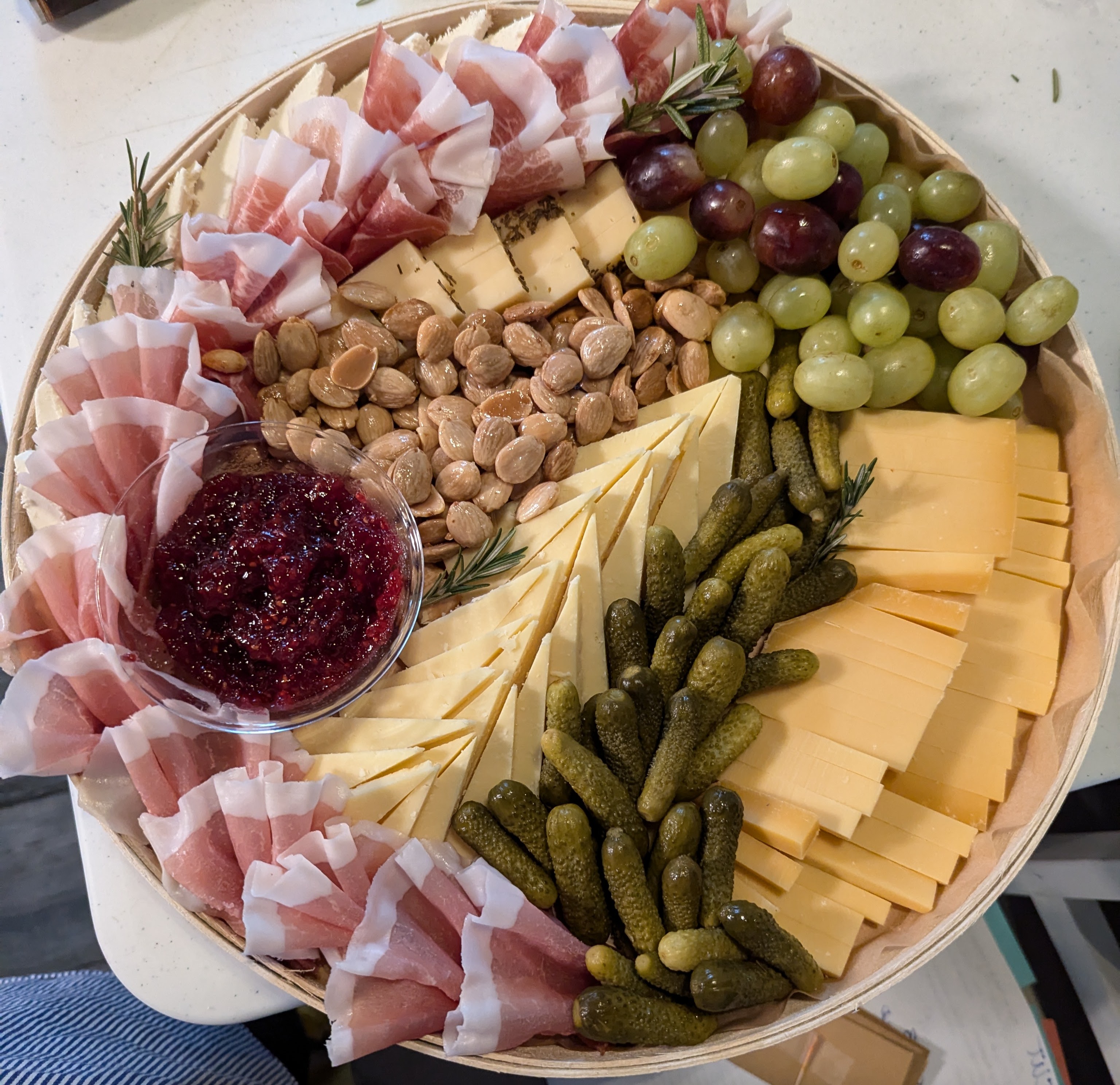 A round wooden board piled with sliced cheeses, prosciutto, grapes, almonds, cornichons, and berry jam.