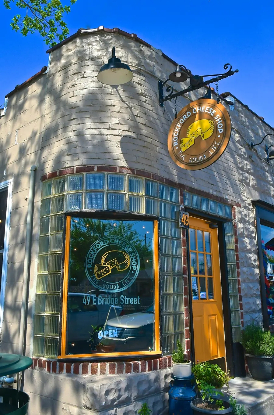 The Rockford Cheese Shop storefront at golden hour — blue glass-block facade, yellow door, and the round hanging sign.